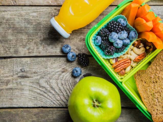 Plastic lunch box with fruits, nuts, carrots, juice, and apple, and sandwich on a wooden table